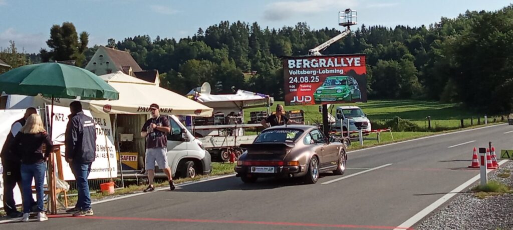 LED-Wand am Start der Bergrallye Voitsberg-Lobming – mobile Outdoor-Anzeige mit 24.08.25 (Training 9:00, Rennen 13:00); braunes Sportcoupé an der Startlinie, Helfer und Zelte.